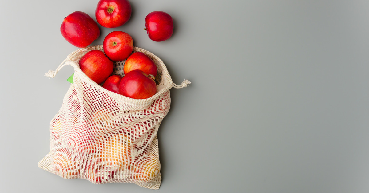 A close-up of several red apples spilling out of a mesh cotton produce bag against a light gray background.