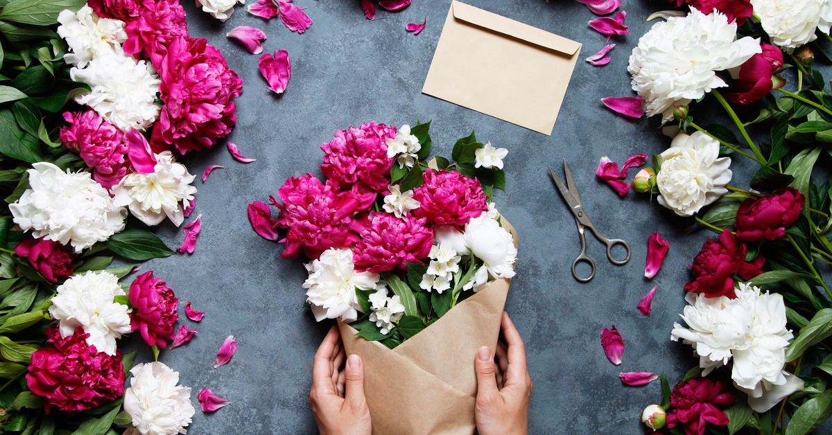 Someone’s hands holding a bouquet of pink and white flowers wrapped in kraft paper on a table full of flowers.