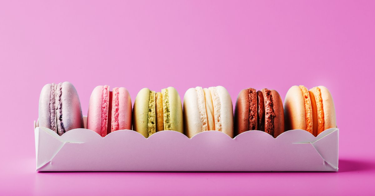 Closeup of six colorful macarons sitting inside an eco-friendly white packing container against a light violet background.