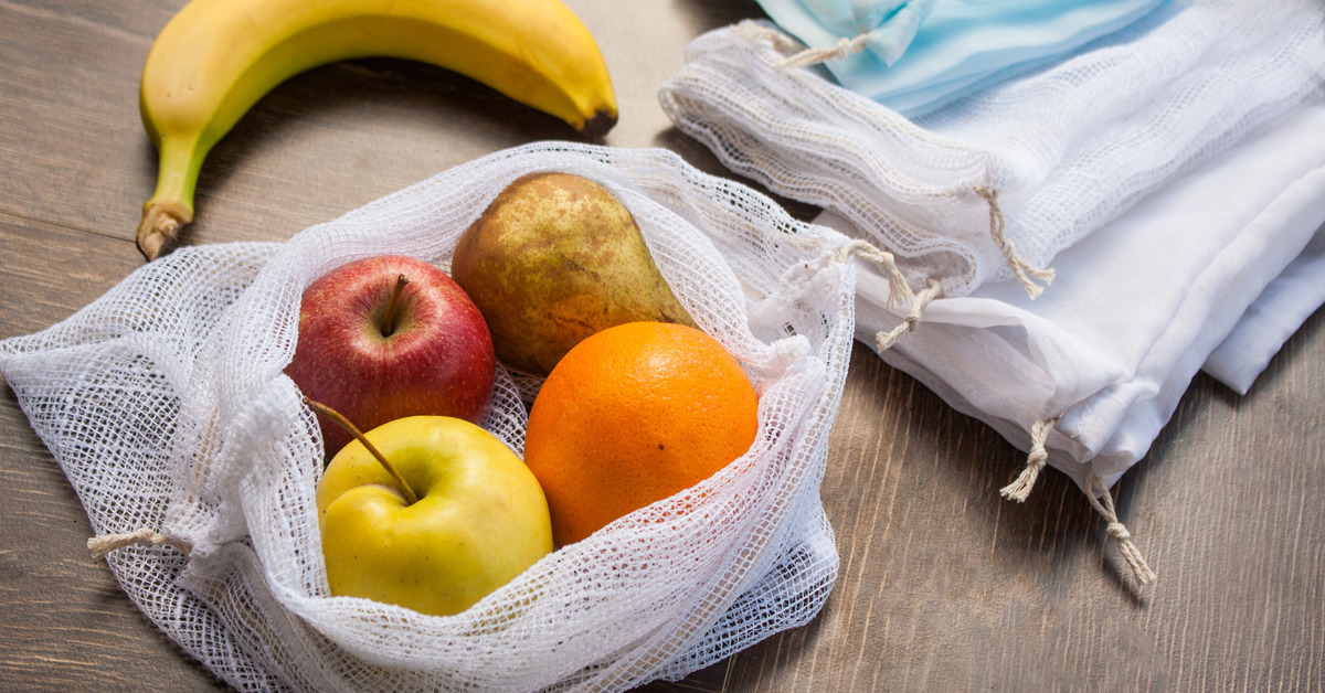 Close-up of a stack of mesh produce bags on a table sitting next to an open bag filled with apples, pears, and oranges.