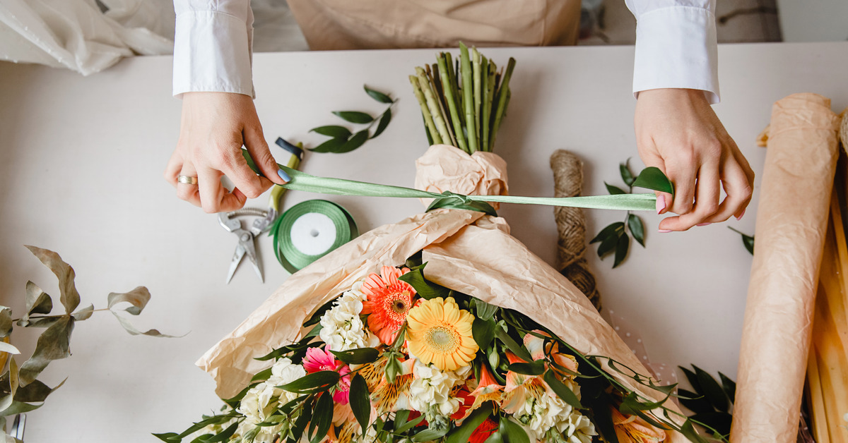 A female florist ties a green ribbon around the stems of a bouquet of flowers wrapped in brown kraft paper.