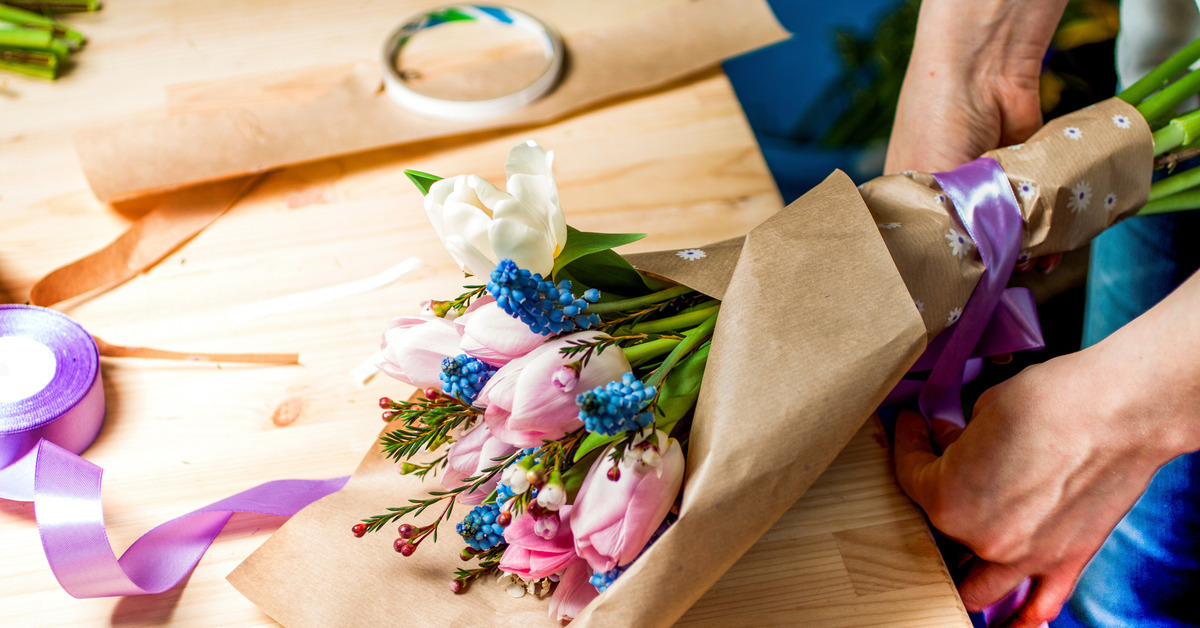 Closeup of someone’s hands wrapping kraft paper and a blue ribbon around a bouquet of tulips on a wood table.