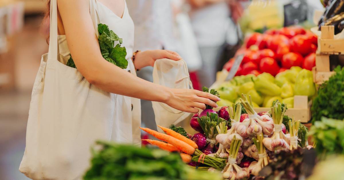 A close-up of a woman shopping at a farmer’s market while filling reusable, eco-friendly bags with fresh produce.