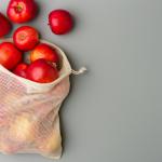 A close-up of several red apples spilling out of a mesh cotton produce bag against a light gray background.